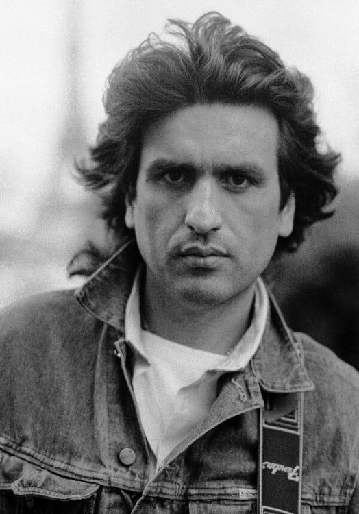 A black-and-white portrait of Toto Cutugno as a young man, in a jeans jacket with a Fender guitar strap.